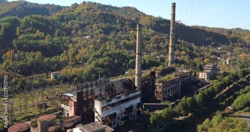  aerial around smooth view ruined factory with high chimneys in postwar abandoned town among picturesque forestry hills under blue sky