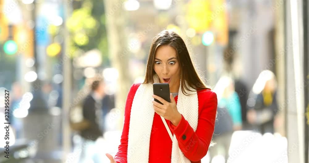 Front view portrait of a shocked woman walking towards camera reading online news on a smart phone in the street Front view portrait of a shocked woman walking towards camera reading online news on a smart phone in the street