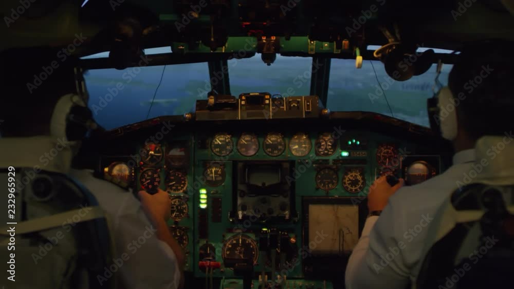 Medium shot from inside of cockpit: male pilots in headsets pressing switches on instrument Panel and controlling airplane with yokes