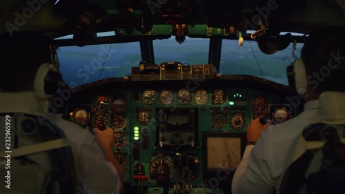 Medium shot from inside of cockpit: male pilots in headsets pressing switches on instrument Panel and controlling airplane with yokes photo