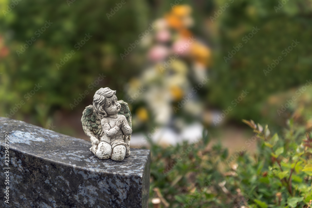 An angel figure sits on a tombstone. In the background flowers on a grave after burial. Concept: sadness or death An angel figure sits on a tombstone. In the background flowers on a grave after burial. Concept: sadness or death