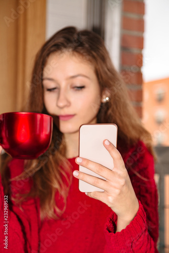 lovely girl in sweater with cup of beverage and mobile phone near window
