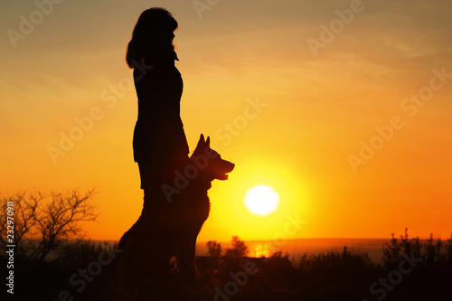 silhouette woman walking with a dog enjoying the sun set over the horizon in the field, pet sitting near girl's leg on nature, concept friendship human and animal