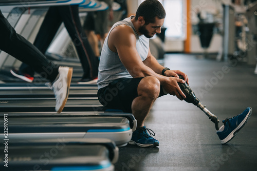 side view of exhausted young sportsman with artificial leg sitting on treadmill at gym photo