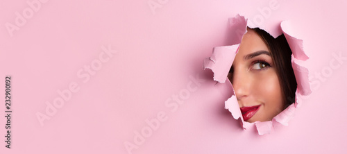 Beauty salon advertising banner with copy space. Beautiful girl looks through hole in pink paper background. Make up artist, fashion, beauty concept. Cosmetics sale.