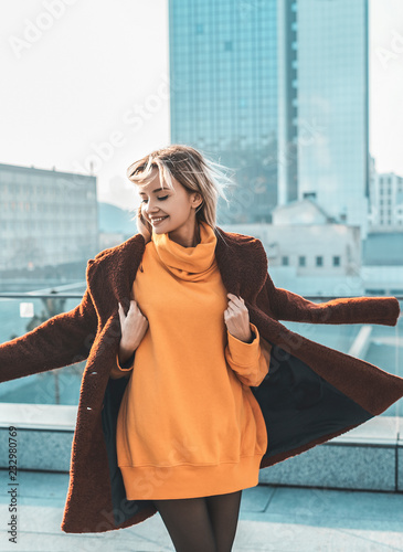  A beautiful young girl blonde walks through the streets of the city, smiling, waving her coat. She is wearing an orange sweater and black tights, a red coat. Street casual style. Emotion of joy