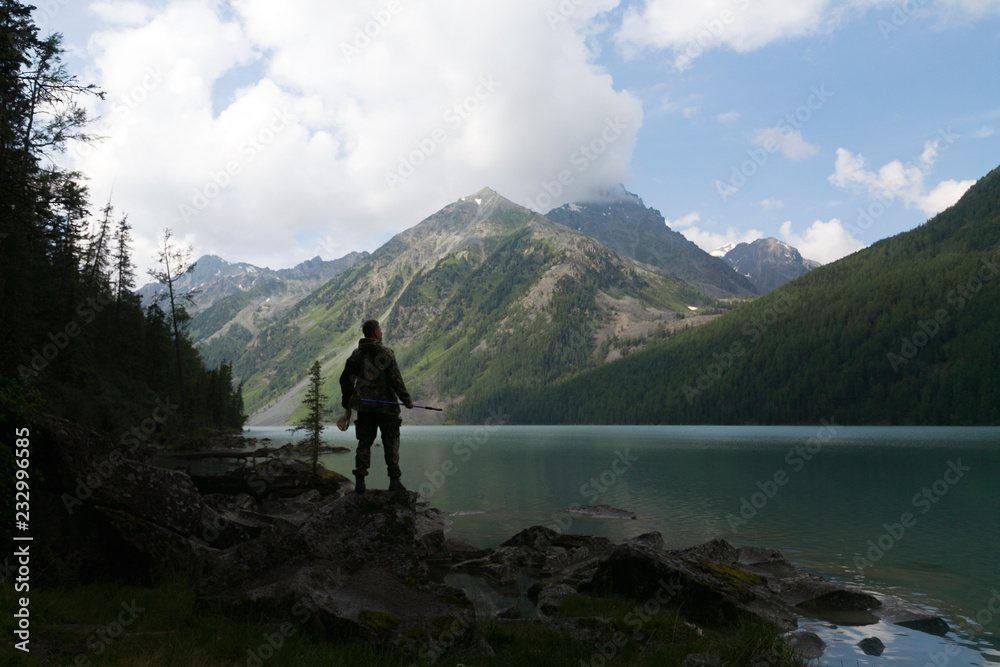 Silhouette of a male tourist against the background of a lake and mountains. A man tourist stands on the shore of a beautiful mountain lake against the backdrop of high mountains.