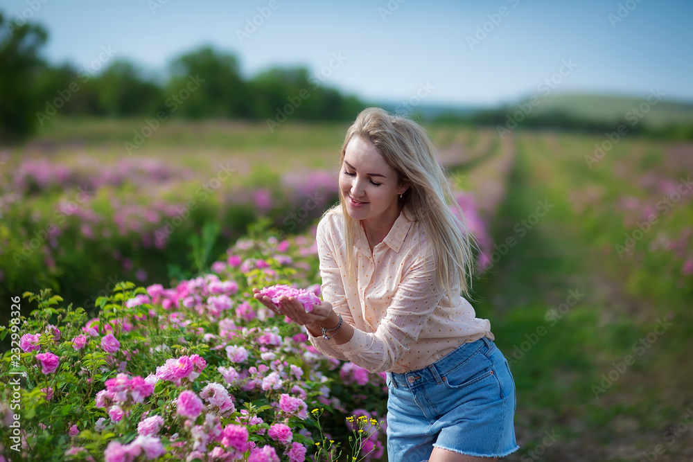 Pretty Young tender woman walking in the tea roses field. Blond lady wearing jeans and retro hat enjoy summer day Pretty Young tender woman walking in the tea roses field. Blond lady wearing jeans and retro hat enjoy summer day