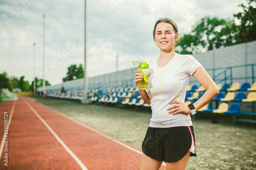 Theme is sport and health. Beautiful young caucasian woman with big breasts athlete runner stands resting on running stadium, running track with bottle in hands drinking water in short shorts © Elizaveta