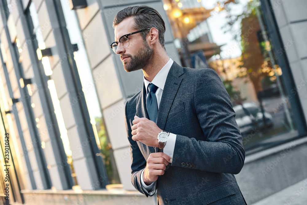 Naklejka premium Close up profile portrait of a successful young bearded guy in suit and glasses. So stylish and nerdy. Outdoors on a sunny street, fixing his cuffs