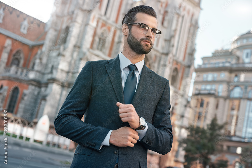 Close up profile portrait of a successful young bearded guy in suit and glasses. So stylish and nerdy. Outdoors on a sunny street, fixing his cuffs