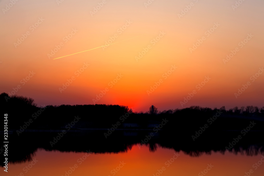 Silhouette of the lake shoreline, symmetrically reflecting the orange yellow sky, the diagonal trace of the aircraft, simple gradient, space for text. Abstract sunset background, Kuskovo park, Moscow.