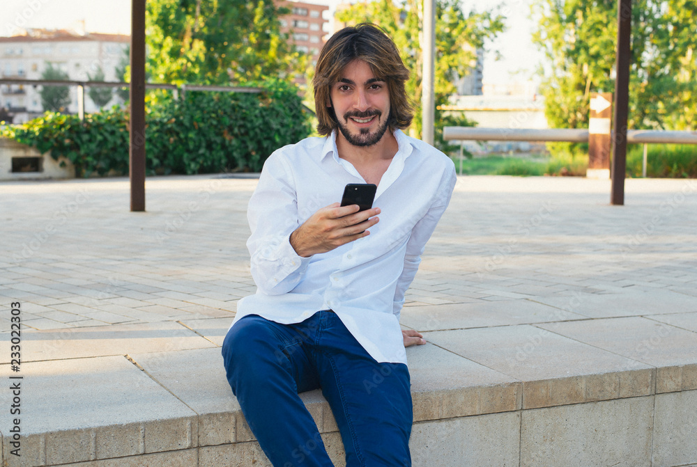 Attractive young man with beard, with white shirt holds a smartphone in his hand and is in the park, smiles. Fashion Attractive young man with beard, with white shirt holds a smartphone in his hand and is in the park, smiles. Fashion