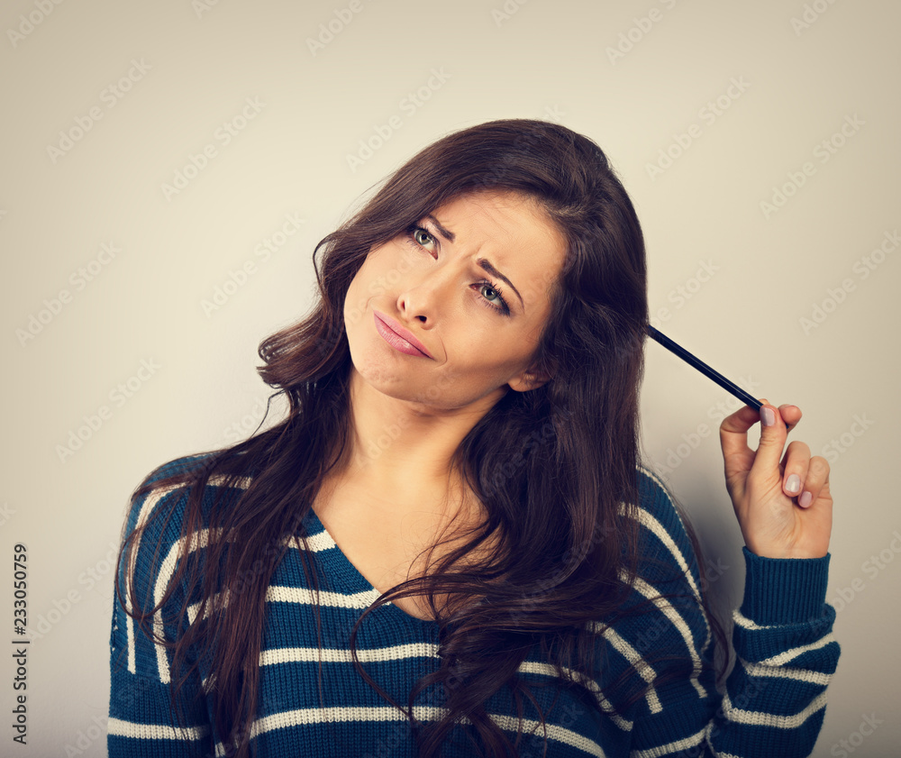 Obraz premium Confusion grimacing brunette woman thinking and looking up with question in eyes and pencil on the head in warm winter sweater. Toned portrait