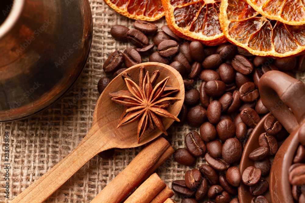 Fototapeta premium Wooden spoon with anise stars and cinnamon lie near the Turk for making Coffee, oranges, and overflowing Mug with coffee beans which are scattered on the table with burlap cloth on. Top view. Close up