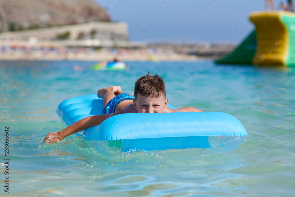European boy in striped shorts is swimming on the blue floater. He is spending his vacations on Canarias.
