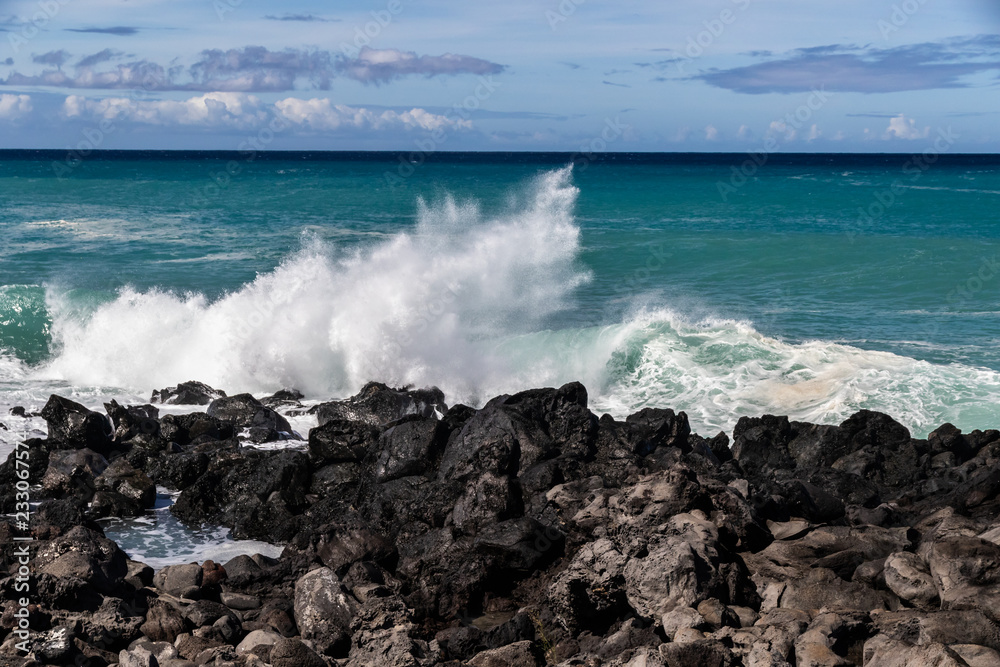 Fototapeta premium Wave breaking on the western Kona coast of Hawaii's Big Island near South Point. White sea spray thrown into the air; Deep blue-green Pacific ocean, and blue sky with clouds in the background. 