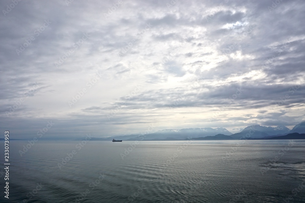 Cook Inlet, Alaska, USA: A freighter on the horizon  against mountains shrouded in morning mist, with dense, dramatic clouds overhead.