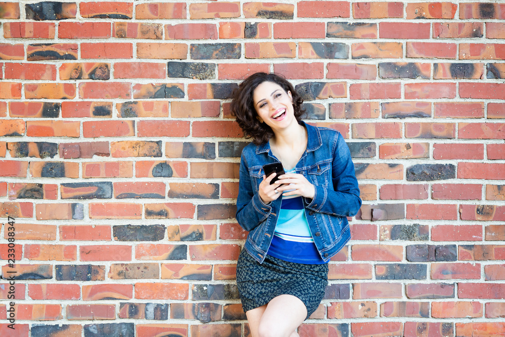 A young Caucasian woman in blue jeans jacket against red brick wall laughing,  holds black smartphone