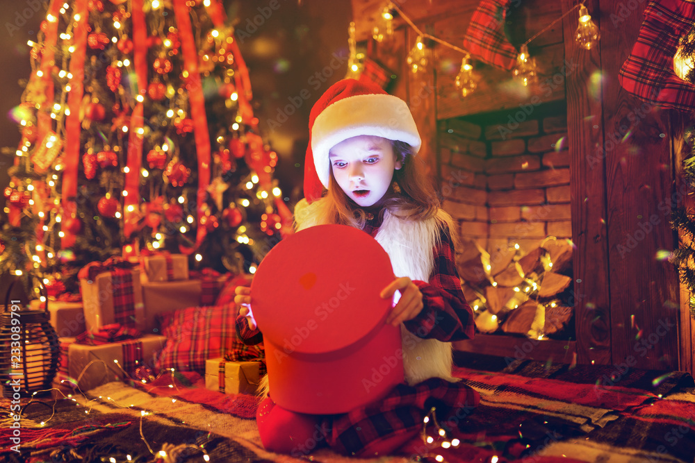 Adorable little girl in santa hat opening a magical Christmas gift by a Christmas tree in cozy living room in winter. Snow frame added. New Year card Adorable little girl in santa hat opening a magical Christmas gift by a Christmas tree in cozy living room in winter. Snow frame added. New Year card