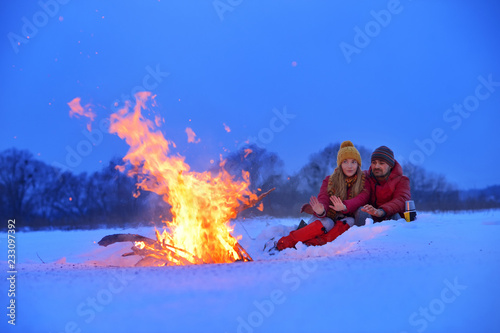 Happy loving couple sitting at a fire in the snow among the fields in the winter against the background of the forest and the sky on Valentine's Day.