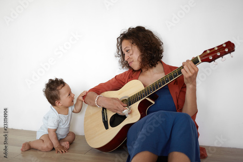 Picture of cute barefooted one year old infant boy sitting on floor with his positive young mom who is playing guitar, singing his favorite lullaby. People, music, children and entertainment photo