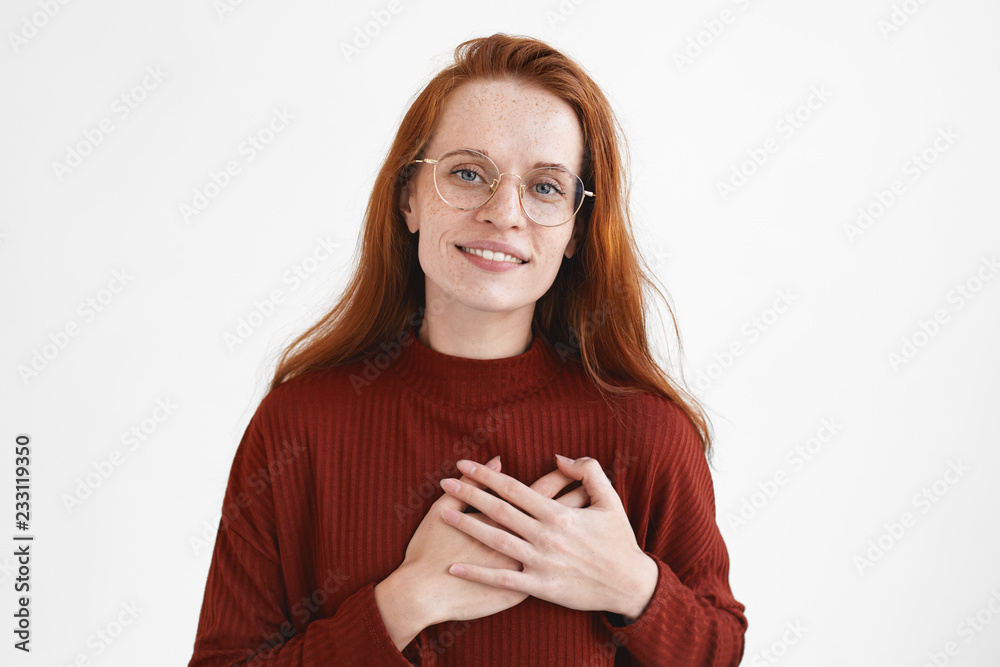 Adorable ginger girl in eyewear keeping both hands on her chest and smiling joyfully, looking with love and gratitude at camera, expressing thankfulness to friend for help and advice, being grateful Adorable ginger girl in eyewear keeping both hands on her chest and smiling joyfully, looking with love and gratitude at camera, expressing thankfulness to friend for help and advice, being grateful