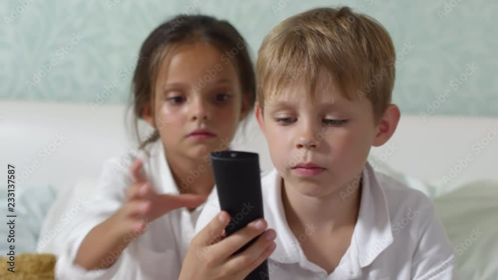 Blonde little boy and his sister sitting on bed and using remote control while watching TV together Blonde little boy and his sister sitting on bed and using remote control while watching TV together