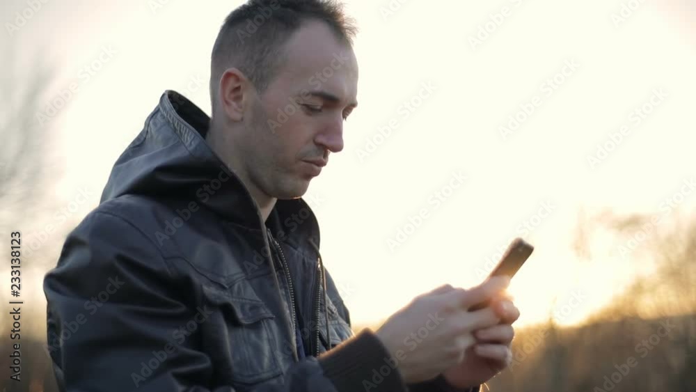 Young smiling man texting message on smartphone outdoors against sunset, slowmotion Young smiling man texting message on smartphone outdoors against sunset, slowmotion