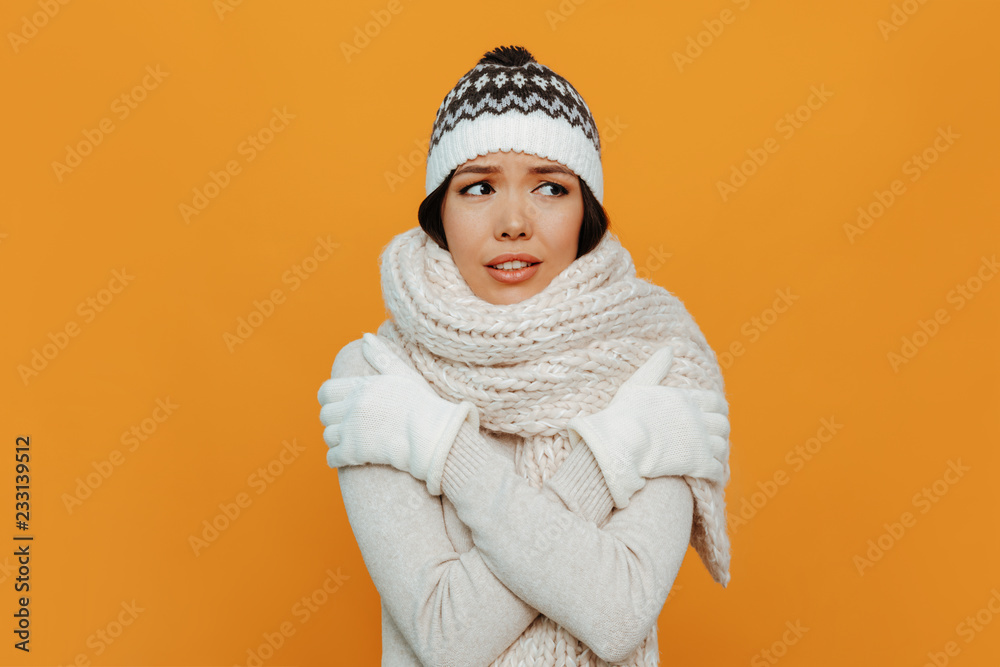 Woman portrait. Accessories. Warmness. Asian girl in a white scarf, cap and gloves is showing she is cold, on an orange background Woman portrait. Accessories. Warmness. Asian girl in a white scarf, cap and gloves is showing she is cold, on an orange background