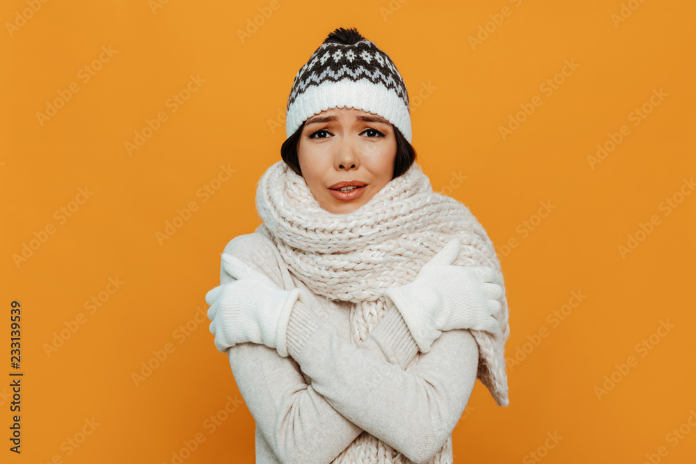 Woman portrait. Accessories. Warmness. Asian girl in a white scarf, cap and gloves is showing she is cold, on an orange background