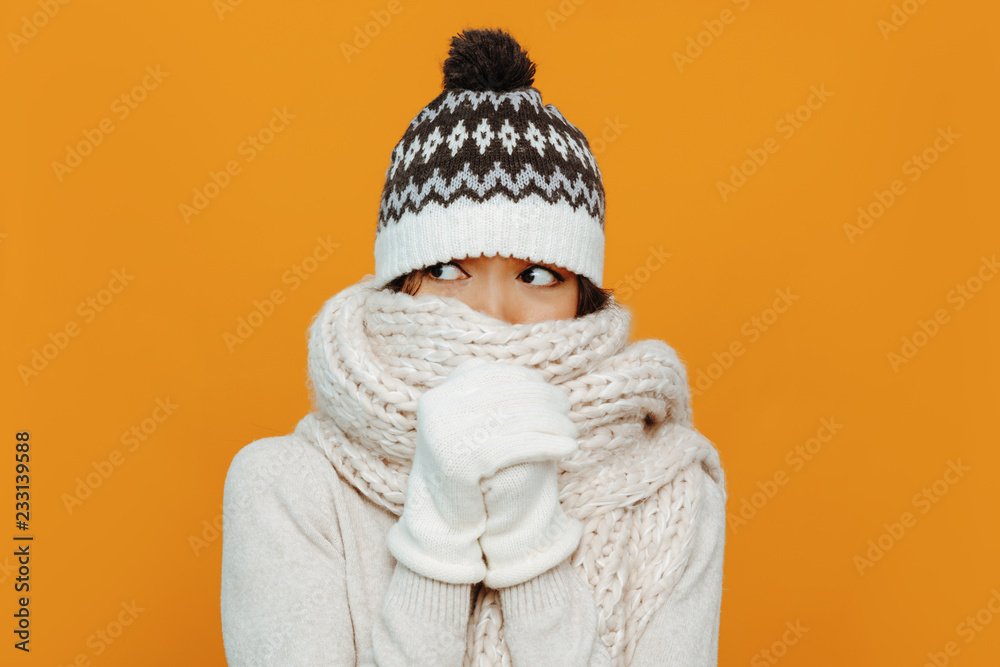 Woman portrait. Accessories. Warmness. Asian girl in a white scarf, cap and gloves is showing she is cold, on an orange background Woman portrait. Accessories. Warmness. Asian girl in a white scarf, cap and gloves is showing she is cold, on an orange background