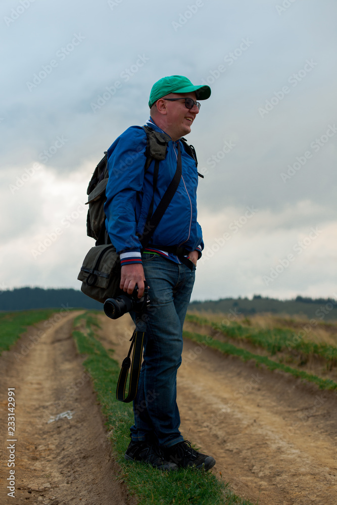 photographer in blue windbreaker and jeans with a camera and a backpack is standing on a dirt road in the mountains