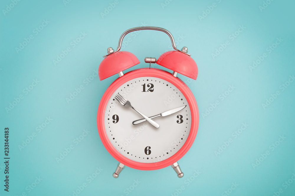 Alarm clock. Fork and knife instead of clock hands. Concept of intermittent fasting, lunchtime, diet and weight loss Alarm clock. Fork and knife instead of clock hands. Concept of intermittent fasting, lunchtime, diet and weight loss