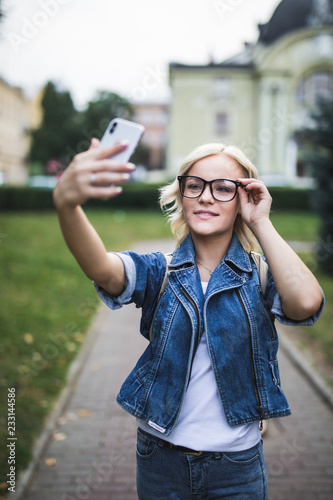 Young attractive woman is making selfie wearing glasses and backpack on the phone outdoors © dianagrytsku