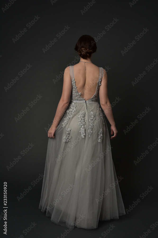 full length portrait of brunette girl wearing beautiful long gown, standing pose with background to the camera on grey studio background. full length portrait of brunette girl wearing beautiful long gown, standing pose with background to the camera on grey studio background.