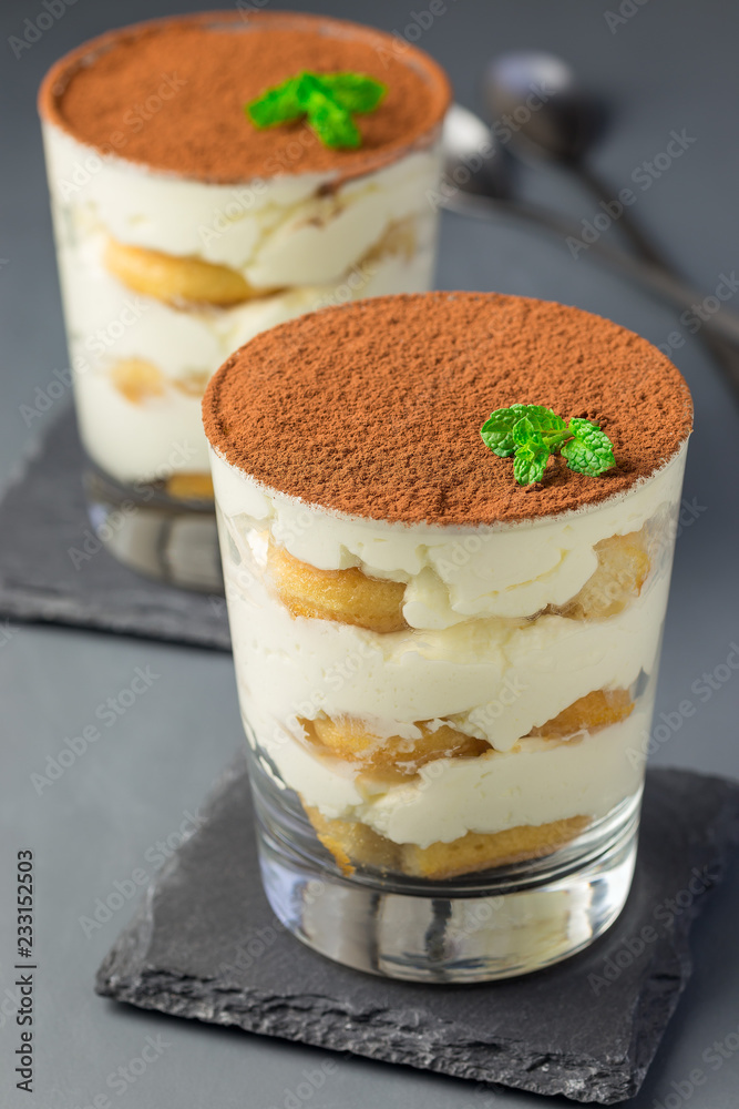 Traditional italian Tiramisu dessert cake in glass, decorated with cocoa powder and mint, on a gray background, vertical Traditional italian Tiramisu dessert cake in glass, decorated with cocoa powder and mint, on a gray background, vertical