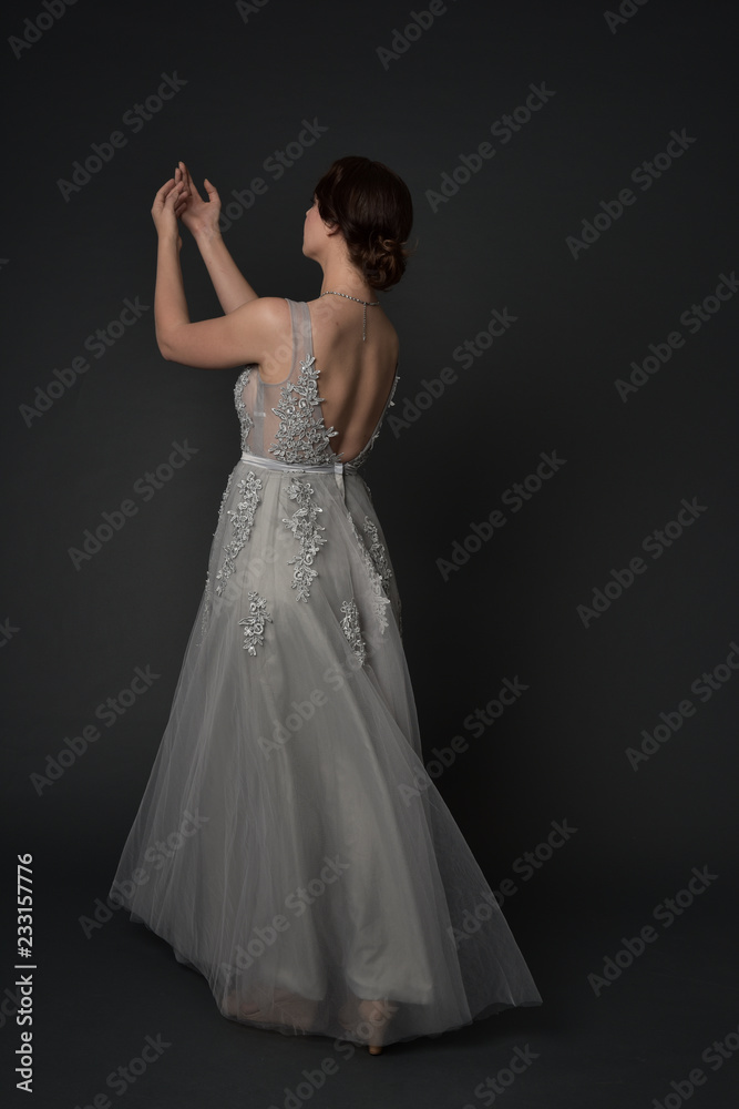 full length portrait of brunette girl wearing beautiful long gown, standing pose with background to the camera on grey studio background. full length portrait of brunette girl wearing beautiful long gown, standing pose with background to the camera on grey studio background.