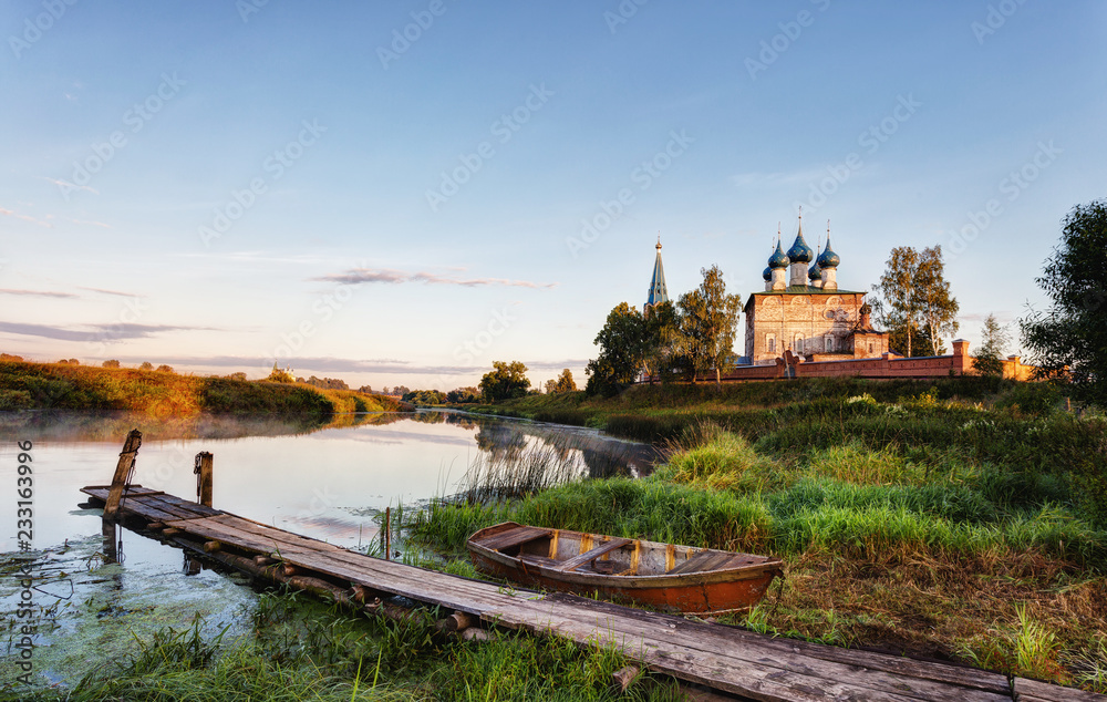 Rural landscape with the wooden bridge, the old boat and church in the early summer morning