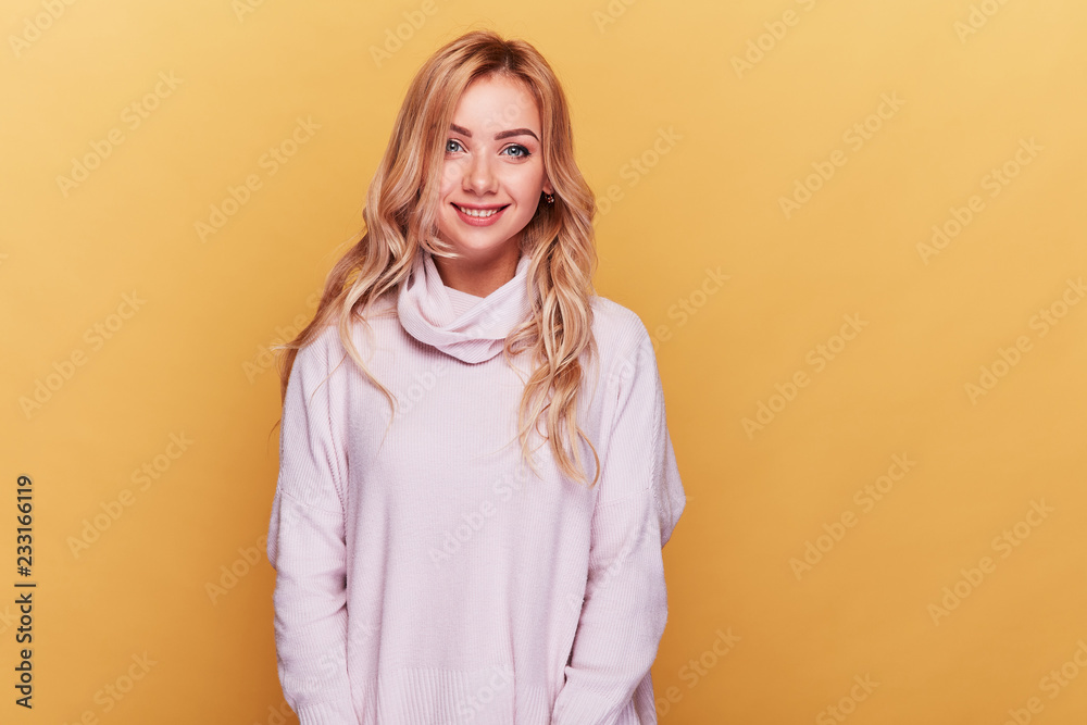 Young cheerful woman with long blonde wavy hair and in light pink sweater standing on yellow background. Young cheerful woman with long blonde wavy hair and in light pink sweater standing on yellow background.