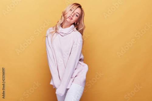 Cute woman with long blond wavy hair dressed in light pink sweater standing on yellow background © monchak
