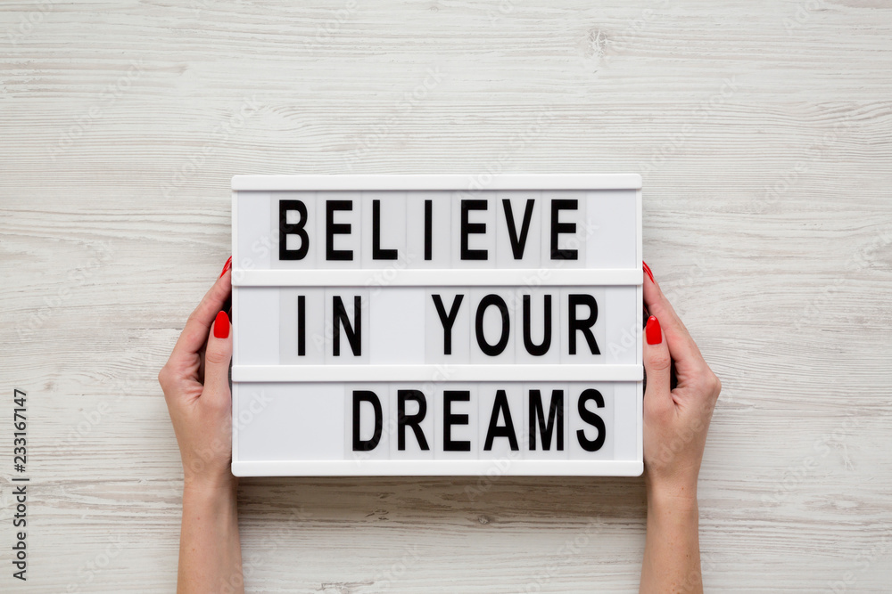 Female hands hold modern board with text 'Believe in your dreams' over white wooden background. From above, overhead, flat-lay. Female hands hold modern board with text 'Believe in your dreams' over white wooden background. From above, overhead, flat-lay.
