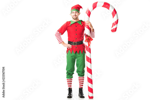 smiling man in christmas elf costume with hand on hips standing near big candy cane isolated on white photo