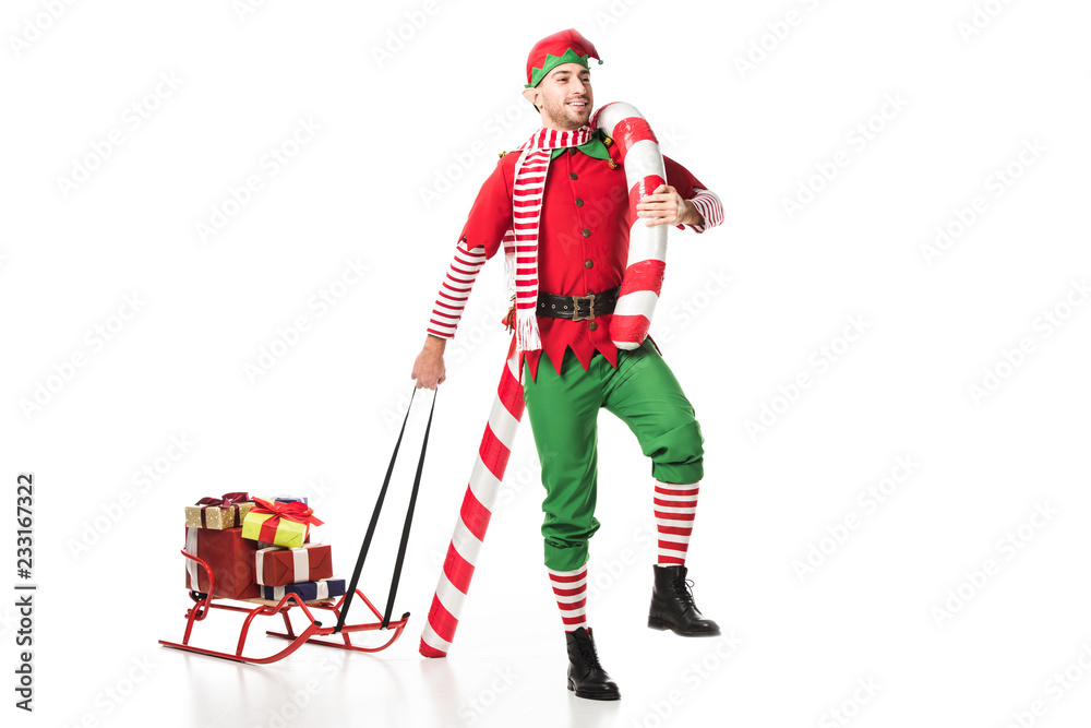 Fototapeta premium smiling man in christmas elf costume carrying sleigh with presents and big candy cane isolated on white