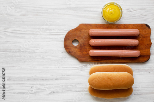Hotdog ingredients: sausages on wooden board, hot-dog buns and mustard on white wooden surface, top view. Flat lay, overhead, from above. Copy space.