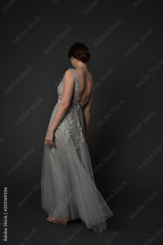 full length portrait of brunette girl wearing beautiful long gown, standing pose with background to the camera on grey studio background. full length portrait of brunette girl wearing beautiful long gown, standing pose with background to the camera on grey studio background.