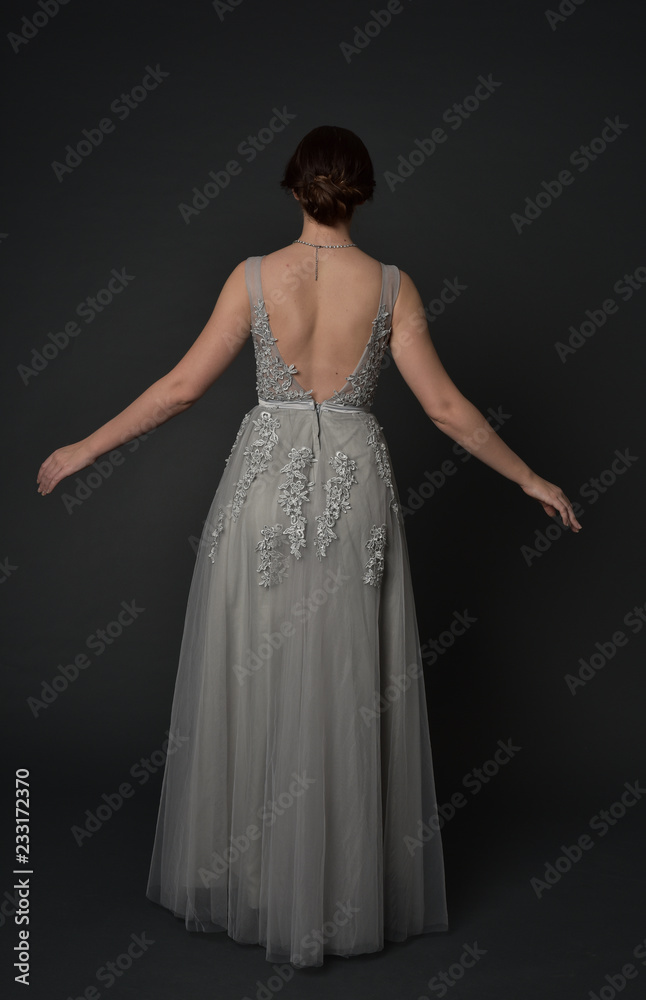 full length portrait of brunette girl wearing beautiful long gown, standing pose with background to the camera on grey studio background. full length portrait of brunette girl wearing beautiful long gown, standing pose with background to the camera on grey studio background.