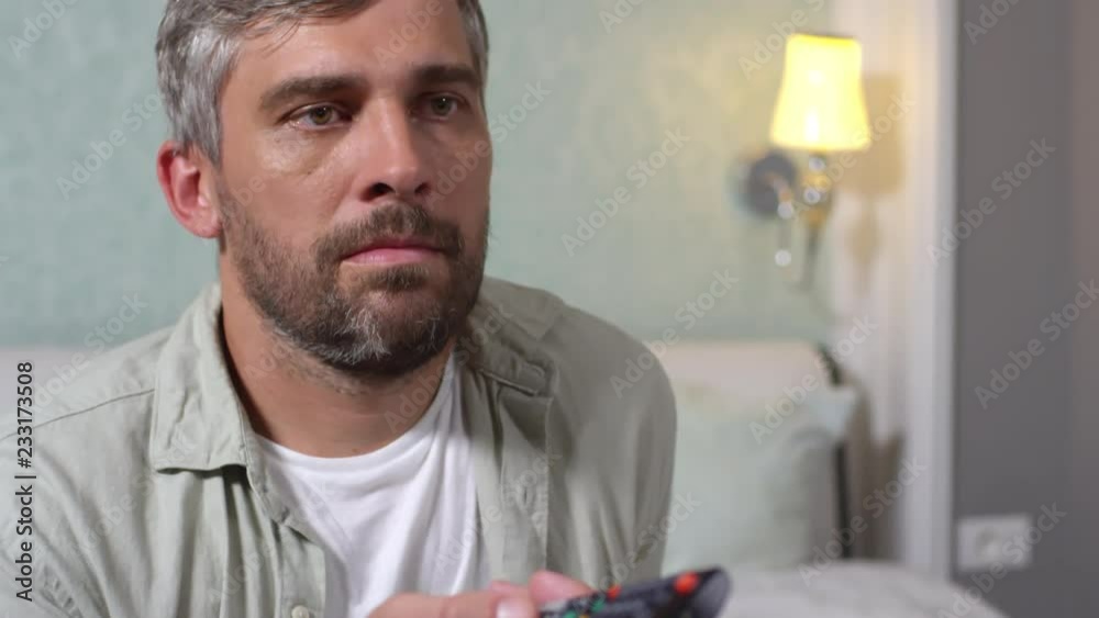Tilt up shot of middle aged bearded man sitting on bed at home and switching TV channels with remote control Tilt up shot of middle aged bearded man sitting on bed at home and switching TV channels with remote control