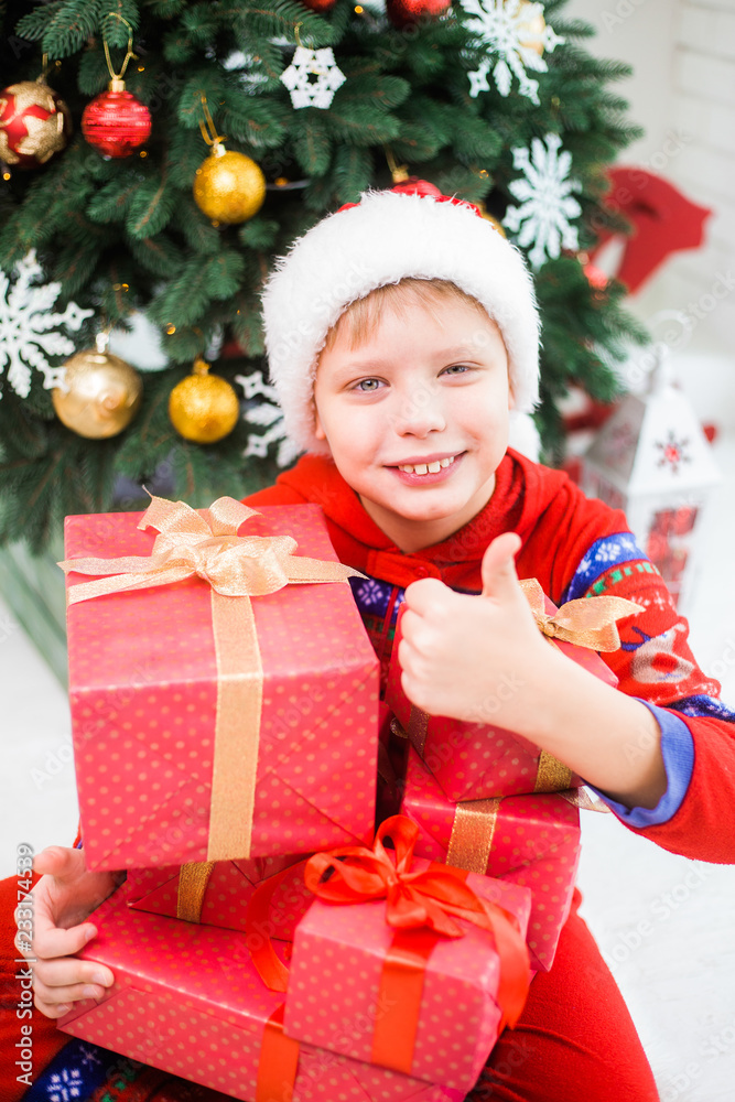 Portrait of cute white kid with thumb up. Boy in red santa hat holding stack of holiday Christmas presents. Vertical color photography.