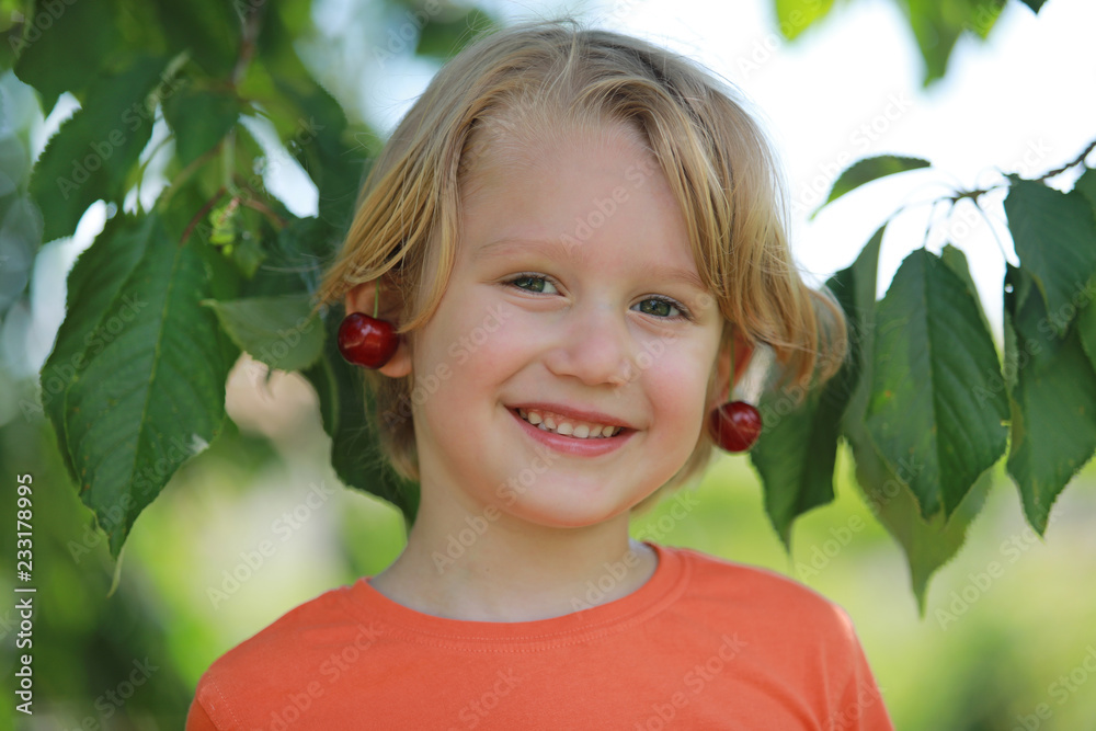 Petit garçon blond avec un grand sourire sous le cerisier avec les cerises sur les oreilles